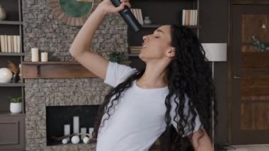 Active energetic young hispanic woman sings rhythmic song using tv remote as microphone sensual dancing to favorite music happy carefree girl dances in room having fun spending free time at home alone