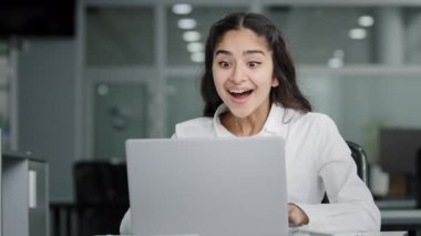 Young excited businesswoman sitting in office looks at laptop screen reading email receiving good news joyful indian girl celebrating success victory surprise about new job opportunity feels pleasure
