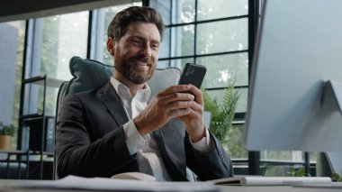 Busy Caucasian bearded man middle-aged 40s businessman uses phone mobile app texting chatting texting message working in office sitting desk with computer remote communicating browsing business chat