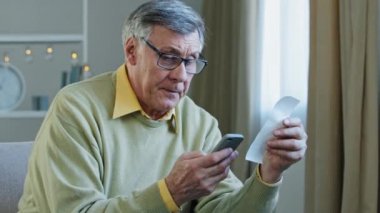 Caucasian 60s talking man mature businessman old householder calculation with calculator app in phone financial check money expense with paper check from bank store e-bank service accounting at home