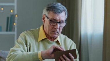 Senior Caucasian 60s man grandpa elderly grandfather sitting at home on couch opening envelope reading letter receive postcard from grandchild smiling win victory gesture success lucky good news offer