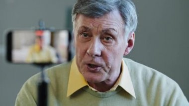 Caucasian old man aged 60s angry negative frustrated influencer blogger at home recording video feedback blog vlog for channel using smartphone on tripod bad news problems speaking into mobile phone