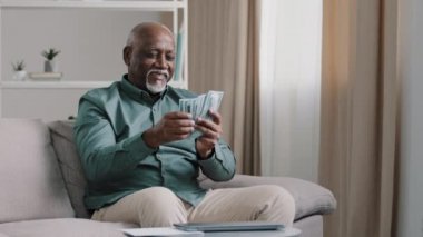 Happy old bald overweight African American senior mature elderly man happy with financial savings salary at home sitting couch. Winning online lottery holding dollars throw up cash money rain falling