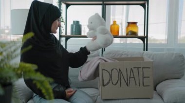 Islamic Muslim woman in black hijab female volunteer help children victims of third world war in Ukraine natural disasters needy poor people sitting on couch packing donations box with clothes and toy