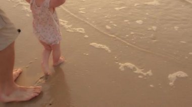 Close up male child barefoot legs stepping in wet water ocean sea play with waves. Unrecognizable father holding baby daughter toddler by hand helping support kid in first steps walking on sand beach