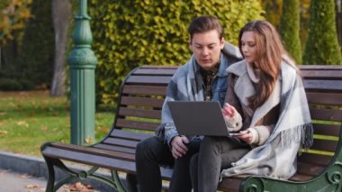 Couple freelancers two partners people young caucasian woman and man entrepreneurs sit on bench in park use urban wifi work on laptop look at device screen make internet order discuss business startup