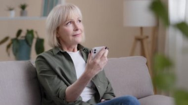 Old Caucasian woman 60s elderly mature lady sits at home on sofa with mobile phone reads news receives notice thinks dreams ponders idea looking away deep in thoughts thinking problem with modern tech