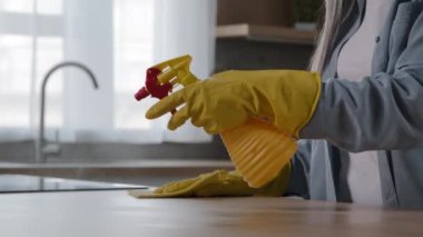 Unknown elderly grey-haired housewife mistress female cleaner housekeeping worker washing kitchen table by wet rag in yellow gloves wiping wooden surfaces at home using detergent antibacterial sprayer