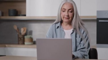 Mature trendy caucasian old businesswoman domestic teacher elderly housewife granny typing on keyboard laptop in kitchen room use home internet check email chatting using service net app browsing news