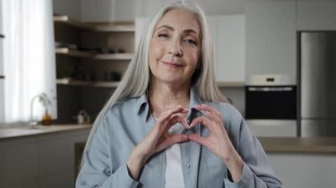 Kindly thankful 60s woman elderly mature volunteer female pensioner gray-haired granny sweetheart makes romantic love frame by fingers heart form valentine symbol expressing human affection tenderness