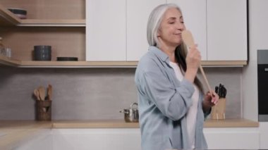 Active grey-haired old woman using wooden spoon like microphone karaoke sing favorite song on domestic kitchen dancing to energetic pop music classic audio track enjoy feel carefree while cook at home