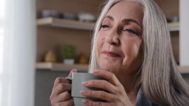 Dreamy inspired old blonde grey-haired woman granny grandmother mature middle aged woman pensioner sitting in kitchen drinking coffee tea hot drink healthy liquid from cup daydreaming relaxing at home