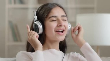 Young teenager carefree arab woman student schoolgirl singer listen favorite music in earphones big headphones active moves enjoy rhythmic musical soundtrack audio song in mp3 playlist resting at home