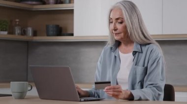 Happy old female caucasian consumer grey-haired granny shopper pay internet order on laptop sits in kitchen hold banking card use online banking app donating money deposit rejoice approved credit loan