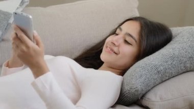 Exhausted latino arab restful lazy young girl woman student housewife fall down on cozy sofa after hard work feel comfort reading sms on smartphone cell gadget chatting with boyfriend shopping online