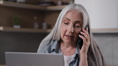 Aged business lady experienced female specialist talk mobile phone look at laptop screen negotiate with client in home kitchen. Grey-haired housewife woman distance communicating use modern smartphone