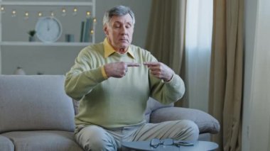 Caucasian old aged funny 60s male grandfather silly male sitting sofa with eyes closed trying to connect two index fingers together checking body coordination doing exercises success then sad failure