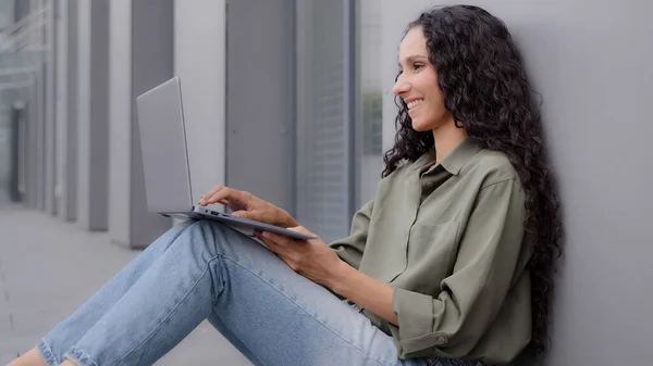 Smiling happy lady with long hair sitting on floor typing browsing laptop casual chatting woman user worker freelancer businesswoman leaning wall outdoors near city building working remote online app