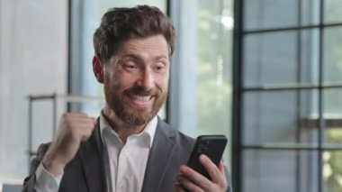 Caucasian bearded businessman 40s boss leader in office with mobile phone victory triumph win victory luck success receive offer money prize opportunity shock ok gesture pointing finger at smartphone
