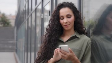 30s lady with curly hair girl standing city near glass wall of company office wall chatting with mobile phone app businesswoman using online net internet service order taxi with smartphone enjoy chat