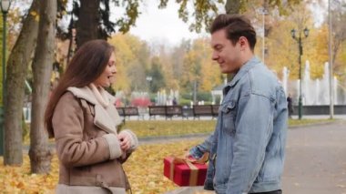 Caucasian boyfriend give red box small gift with gold ribbon to lovely pretty girlfriend young woman in autumn park surprise present for valentines day birthday anniversary symbol of love affection