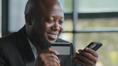 Joyful african shopper consumer american businessman buyer pay bills in mobile app enter security code of credit card in banking app donating make internet order financial operation electronic economy