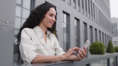 Side view Caucasian Hispanic 30s happy smiling woman standing on balcony terrace in city female businesswoman with phone ecommerce shopping online typing in smartphone chat searching receive message