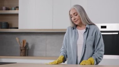 Joyful active older housewife tidy female cleaner domestic maid clean kitchen wooden table by wet in yellow protective gloves wiping interior surfaces washing countertop sanitization daily cleanliness