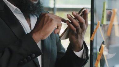 Unknown african bearded man boss specialist american businessman holds mobile phone in office stands near wallboard transparent desk with sticky reminder notes chatting organize corporate online work