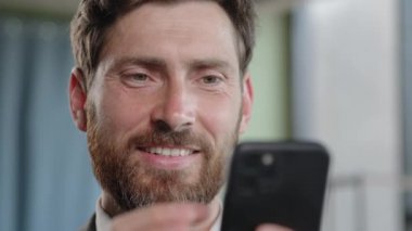 Close up head shot smiling bearded adult 40s businessman holding smartphone in office smile reading good news using online app middle-aged manager with cell phone mobile service texting corporate chat