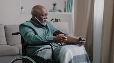 Side view African old 60s male ill senior retired bald man with gray beard measuring blood pressure with electronic medical device and stethoscope sitting in wheelchair at home cardiology healthcare