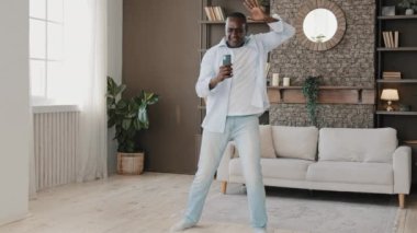 Hilarious energetic senior dancer male artistic african man listen music at home dancing rhythmic to musical track enjoy loud melody playful moving sing favorite song in mobile microphone relax alone