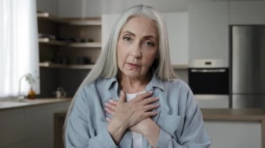 Caucasian old mature woman put folded palms on heart keeps wrinkled hands on chest make thankful trust gesture friendly symbol express sincere feelings smile look at camera believe and charity concept