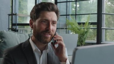 Busy bearded Caucasian adult middle-aged millennial man office worker businessman manager consulting client by telephone reading information on laptop screen looking at computer talk phone answer call