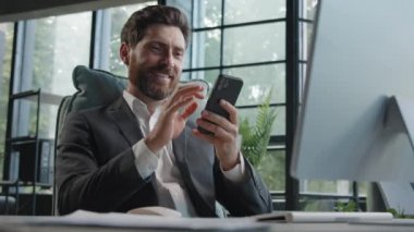 Busy Caucasian bearded man middle-aged 40s businessman uses phone mobile app texting chatting texting message working in office sitting desk with computer remote communicating browsing business chat