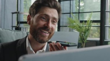 Happy adult 40s male manager bearded businessman sitting at workplace with computer smiling recording audio message sending voicemail using assistant software application on smartphone mobile app chat