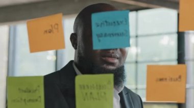 Male worker african businessman read colorful small paper notes on glassy wallboard in office analyzing information stick reminder sticker on transparent board write task list developing strategy plan