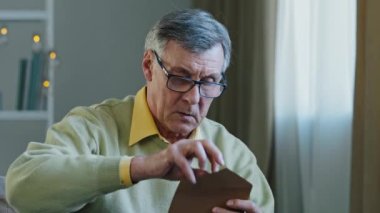 Old senior mature Caucasian 60s man in eyeglasses open envelope notice postcard reads bad news letter reject from bank. Depressed grandfather frustrated about loss of health trouble financial problems