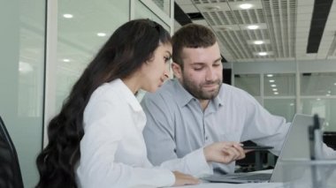 Young man and woman colleagues sitting in office working together at workplace looking at laptop screen creating project discussing planning girl explaining online business service corporate teamwork