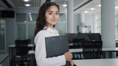 Young confident Indian businesswoman model posing indoors smiling female manager office worker trainee stands holding folder with documents in hand looking at camera advertises business corporation