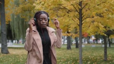 Young funny carefree woman walks in autumn park listens to rhythmic music in headphones dances energetic sings favorite song attractive joyful girl dancer performs dance moves outdoors active moving