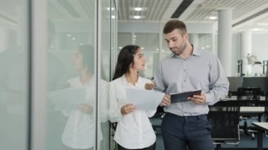 Business young man and woman colleagues stand in office communicate discuss joint project girl trainee asks clarifies details task talks with mentor at workplace creates ideas professional teamwork