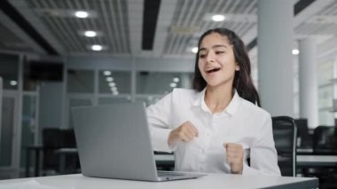 Young happy girl office worker typing on laptop checks email getting electronic message with good news or profitable offer feeling excited dancing dance of winner celebrating success receives reward