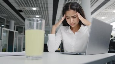 Young tired woman manager sits in office feels headache suffers from pain overwork unhealthy girl office worker feeling unwell migraine pressure syndrome focus on glass of pain medication treatment