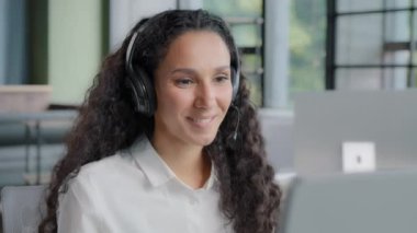 Young positive woman in headphones communicates via video call call center operator talks to customer on online conference via webcam on computer using headset distance office chat e-learning concept