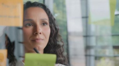 Close-up young pensive businesswoman project organizer writing down new tasks on sticky colored notes developing idea puts reminder creates strategy plans workflow attaching stickers on glass board