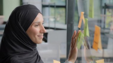 Smiling successful businesswoman corporate leader writing project tasks leaves reminder on sticky colored note organizes work young arab woman office worker in hijab attaches stickers on glass board