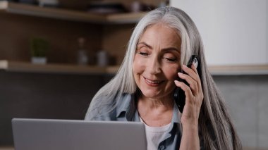 Aged business lady experienced female specialist talk mobile phone look at laptop screen negotiate with client in home kitchen. Grey-haired housewife woman distance communicating use modern smartphone
