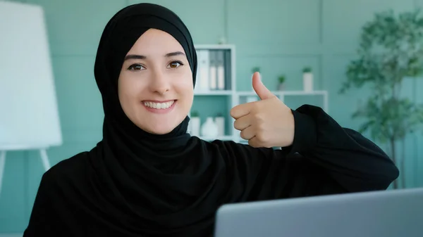 Arabian Islamic Muslim student in black hijab successful businesswoman sitting at workplace home office looking at laptop computer screen reads good news shows like thumbs up agree approve symbol