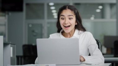 Young woman manager sitting in office looking at laptop screen receiving notification reads good news rejoices in success celebrating victory feels good luck receives promotion at work gets job bonus